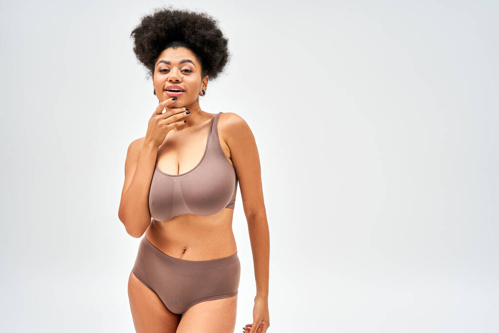 Positive and seductive african american woman in brown panties and bra touching chin and looking at camera while standing  isolated on grey, self-acceptance and body positive concept  - Photo, Image