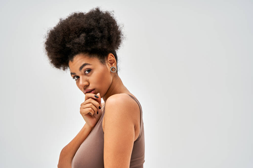 Portrait de femme afro-américaine confiante en soutien-gorge marron moderne touchant le menton et regardant la caméra tout en étant isolé sur le concept gris, l'acceptation de soi et le corps positif  - Photo, image