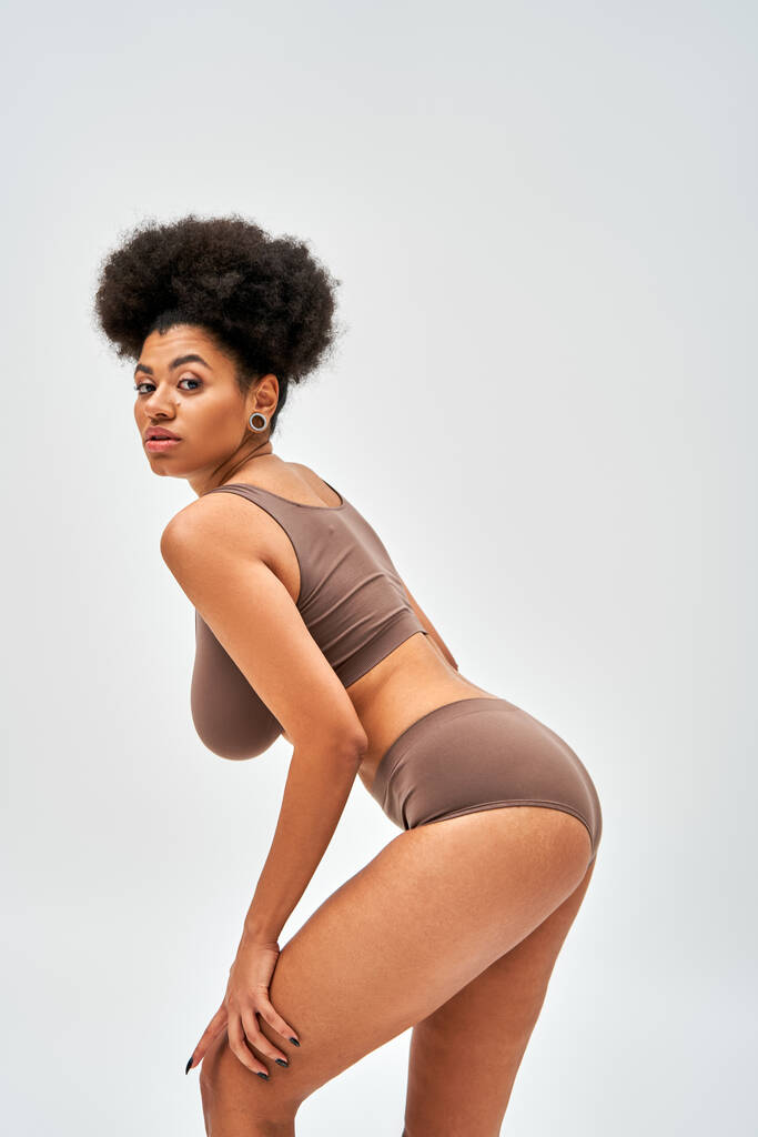 Zelfverzekerde en moderne Afro-Amerikaanse vrouw in bruine lingerie op zoek naar camera tijdens het aanraken van been en poseren geïsoleerd op grijs, zelf-acceptatie en lichaam positief concept  - Foto, afbeelding