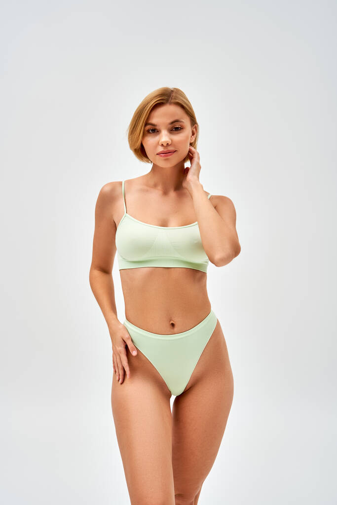 Pretty young blonde woman with natural makeup in light green lingerie touching hip while standing and looking at camera isolated on grey, self-acceptance and body positive concept  - Photo, Image