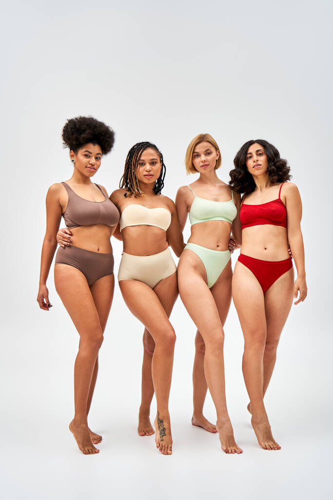 Comprimento total de mulheres multiétnicas sexy e descalça em lingerie colorida abraçando e olhando para a câmera em fundo cinza, modelos multiculturais e conceito de movimento de positividade corporal - Foto, Imagem