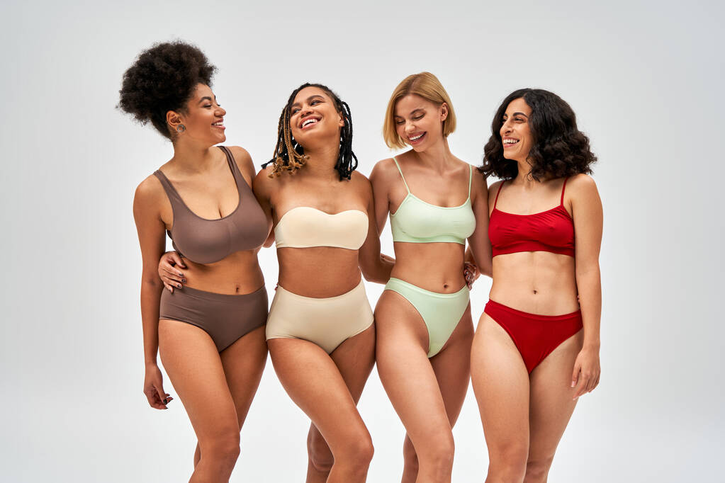 Pozitív és többnemzetiségű magabiztos nők színes fehérnemű ölelésben, miközben egymásra néznek és elszigetelten állnak a szürke, multikulturális modelleken és a test pozitivitásának koncepcióján - Fotó, kép