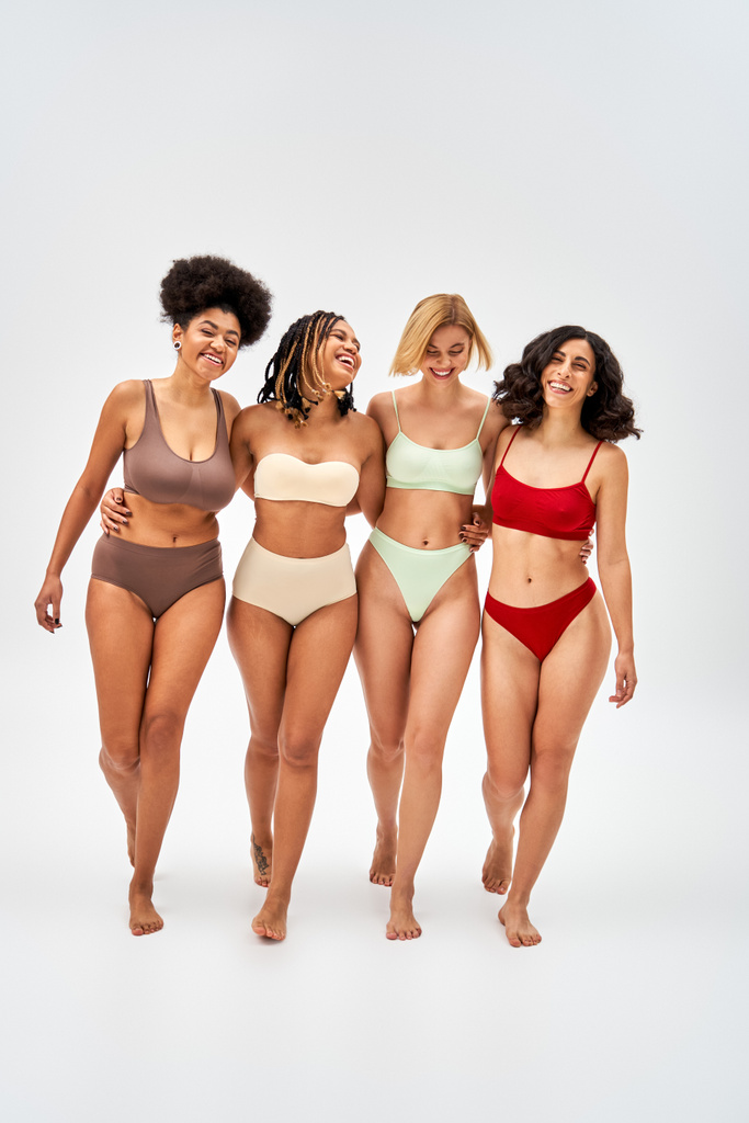 Full length of confident and positive multiethnic women in colorful lingerie hugging and walking on grey background, multicultural models and body positivity movement concept - Photo, Image