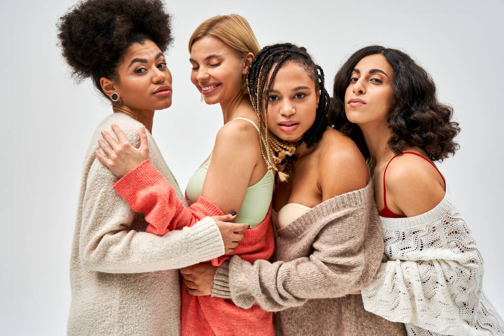 Portrait of group of multiethnic girlfriends in warm knitted jumpers hugging and standing together isolated on grey, different body types and self-acceptance, multicultural representation - Photo, Image