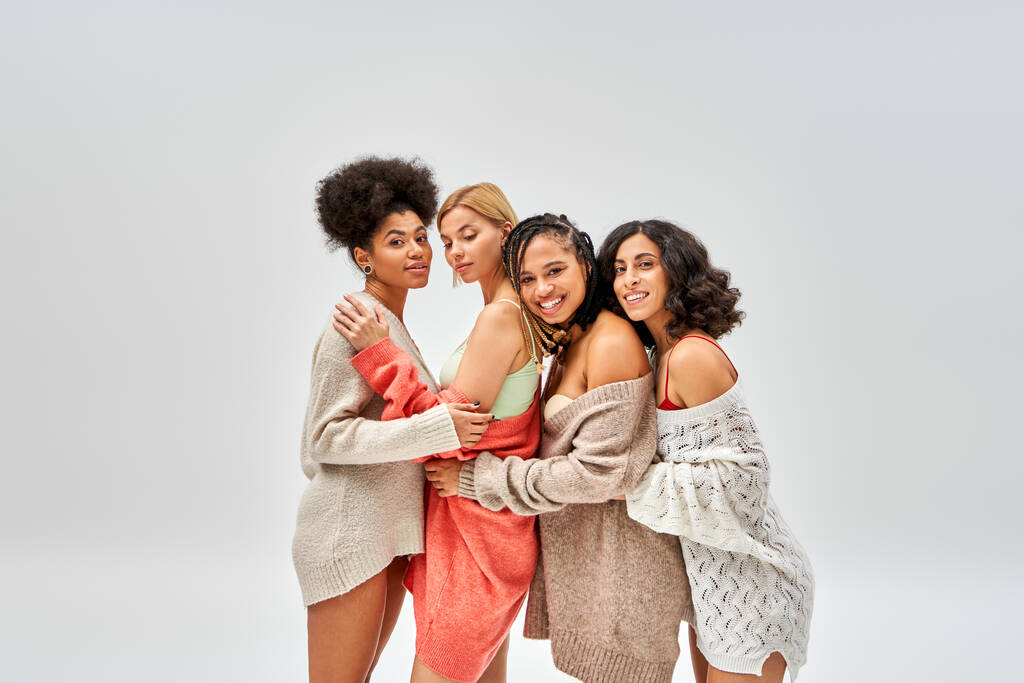 Fröhliche und stylische multiethnische Frauen in Wollpullovern, die sich umarmen und isoliert auf grauen, unterschiedlichen Körpertypen und Selbstakzeptanz stehen, multikulturelle Repräsentation - Foto, Bild