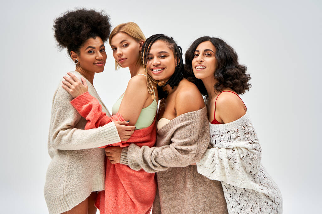 Retrato de mujeres multiétnicas con estilo y confianza en suéteres de punto sonriendo y abrazándose aislados en gris, diferentes tipos de cuerpo y auto-aceptación, representación multicultural - Foto, imagen