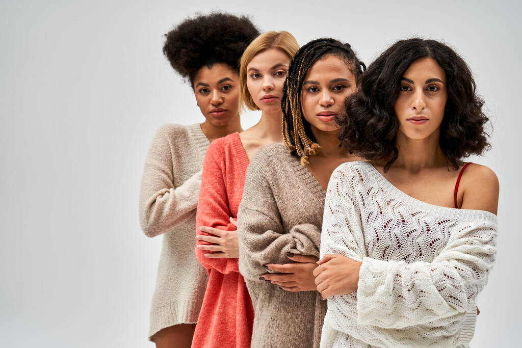 Pewne siebie i wielokulturowe kobiety w stylowych, dzianych swetrach przecinających ramiona i stojące obok siebie na szarych, różnych typach ciała i samoakceptacji, reprezentacji wielokulturowej - Zdjęcie, obraz