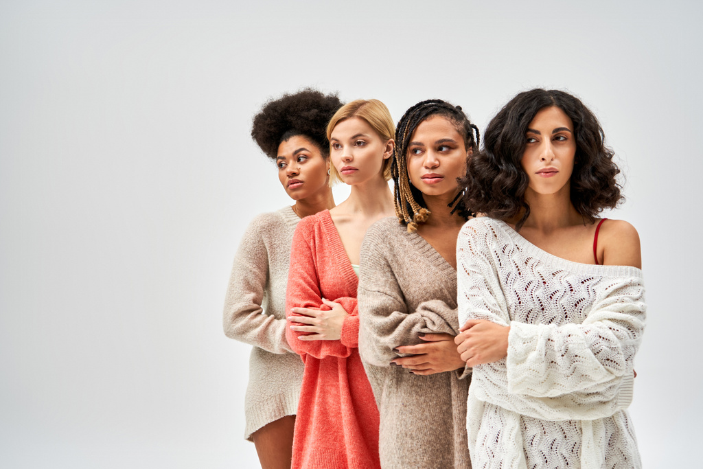 Κομψή πολυεθνική γυναίκες σε μοντέρνα πλεκτά πουλόβερ διασχίζουν τα χέρια και κοιτάζοντας μακριά, ενώ στέκεται απομονωμένο σε γκρι, διαφορετικούς τύπους σώματος και αυτο-αποδοχή, πολυπολιτισμική εκπροσώπηση - Φωτογραφία, εικόνα