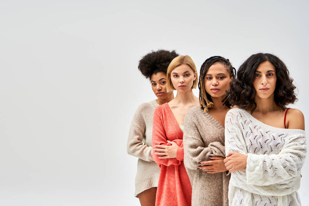 Multietnická skupina žen ve stylových pletených svetrech zkřížených na ramenou a při pohledu na kameru izolovanou na šedé, různé typy těl a sebepřijetí, multikulturní reprezentace, kopírovací prostor - Fotografie, Obrázek