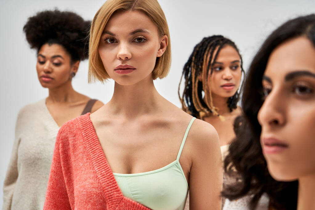 Portrait of young blonde woman in bra and wool jumper looking at camera while standing near blurred friends isolated on grey, different body types and self-acceptance, multicultural representation - Photo, Image