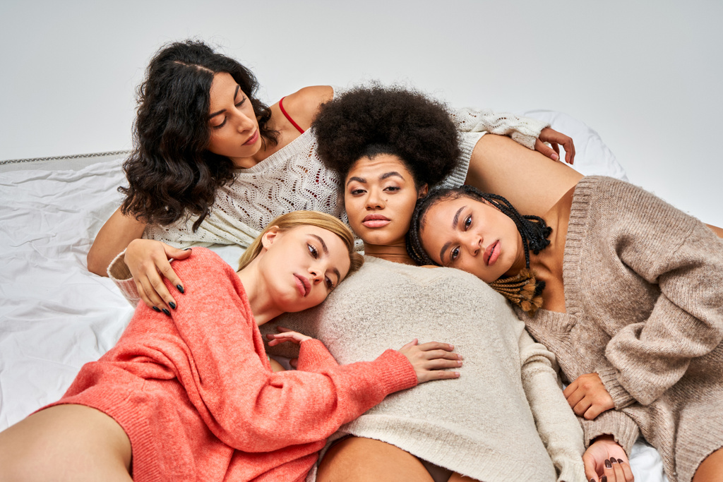 Vista de ángulo alto de las mujeres multiétnicas en elegantes jerséis cálidos mirando a la cámara mientras yacen en la cama aislados en gris, diferentes tipos de cuerpo y auto-aceptación, representación multicultural - Foto, Imagen