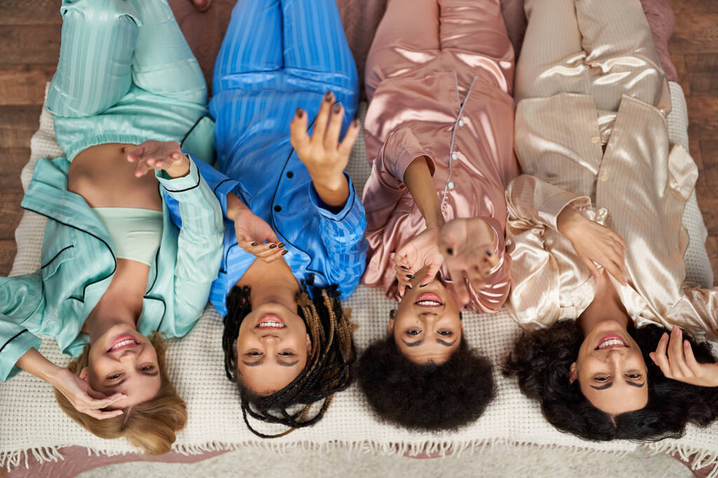 Top view of smiling and multiethnic girlfriends in colorful pajama outstretching hands and looking at camera during pajama party on bed in bedroom at home, bonding time in comfortable sleepwear - Photo, Image