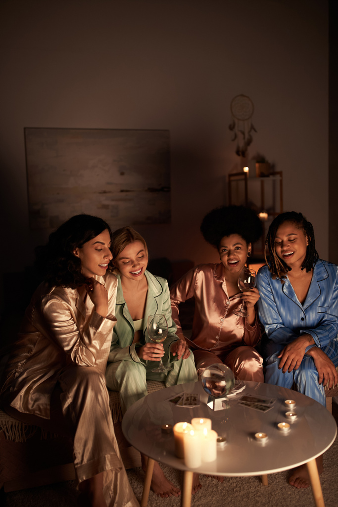 Позитивные многоэтнические девушки в красочной пижаме, держащие бокалы вина возле карт Таро, хрустальный шар и свечи во время девичника дома, время сближения в удобной сонной одежде, гадание  - Фото, изображение