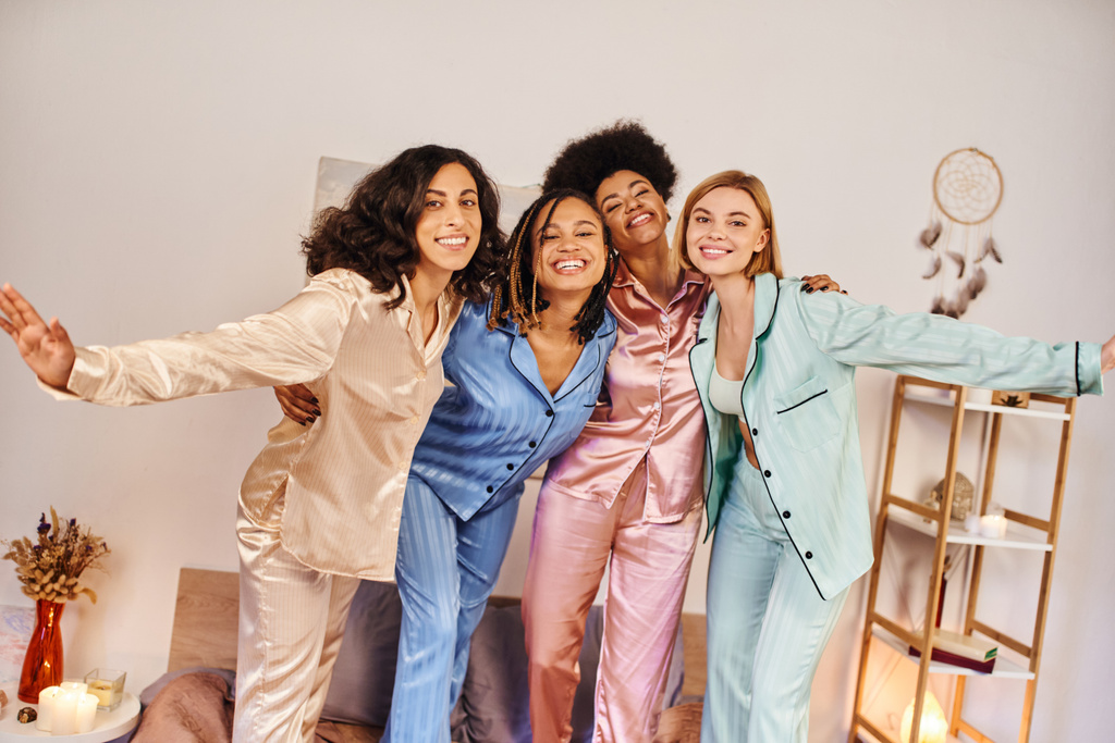 Lächelnde multikulturelle Freundinnen im farbenfrohen Pyjama, die sich umarmen und einander anschauen, während sie zu Hause auf dem Bett stehen und in bequemer Nachtwäsche Zeit miteinander verbringen - Foto, Bild
