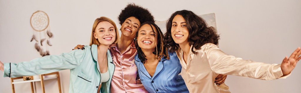 Positive multiethnic friends in colorful pajama hugging each other and looking at camera while having fun at home, bonding time in comfortable sleepwear, slumber party, banner  - Photo, Image