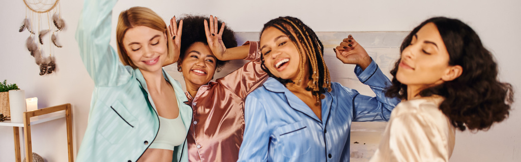 Возбужденные и радостные многонациональные девушки в красочных пижамных танцах и веселятся вместе во время пижамной вечеринки дома, время сближения в удобной сонной одежде, баннер  - Фото, изображение
