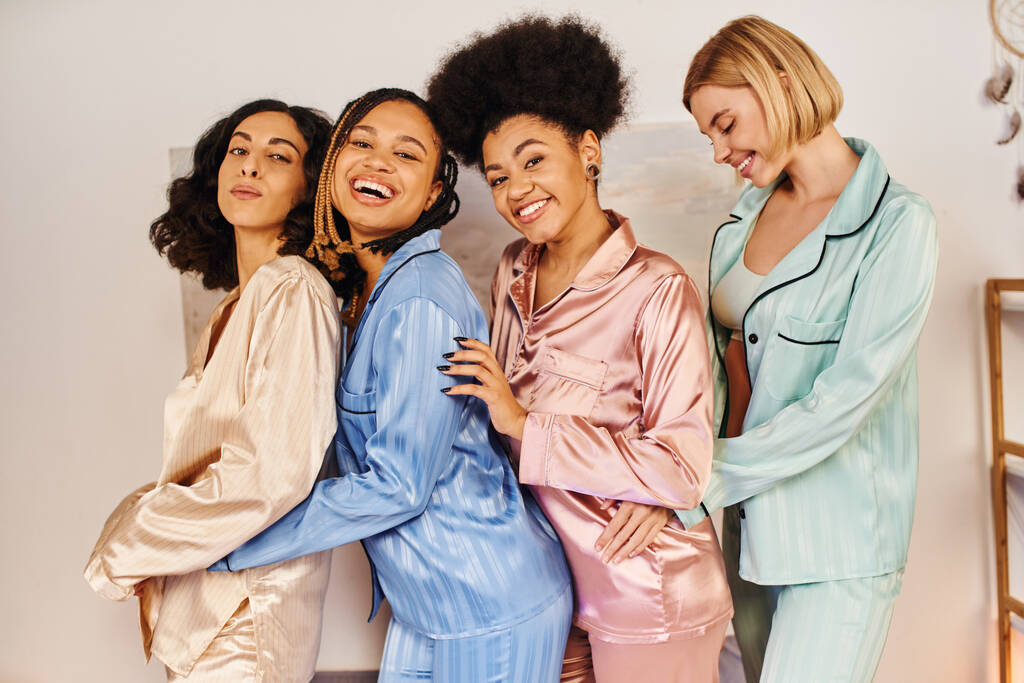 Happy multicultural girlfriends in colorful pajama looking at camera, having fun and hugging each other during pajama party at home, bonding time in comfortable sleepwear, cultural diversity - Photo, Image