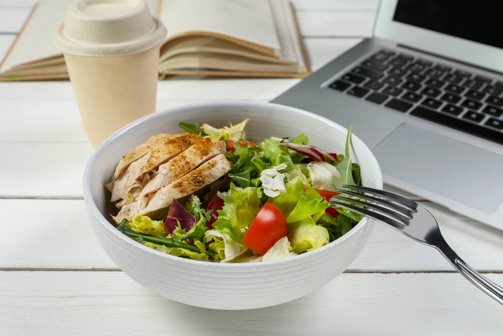 Mísa chutného jídla, vidličky a notebooku na bílém dřevěném stole, detailní záběr. Pracovní oběd - Fotografie, Obrázek