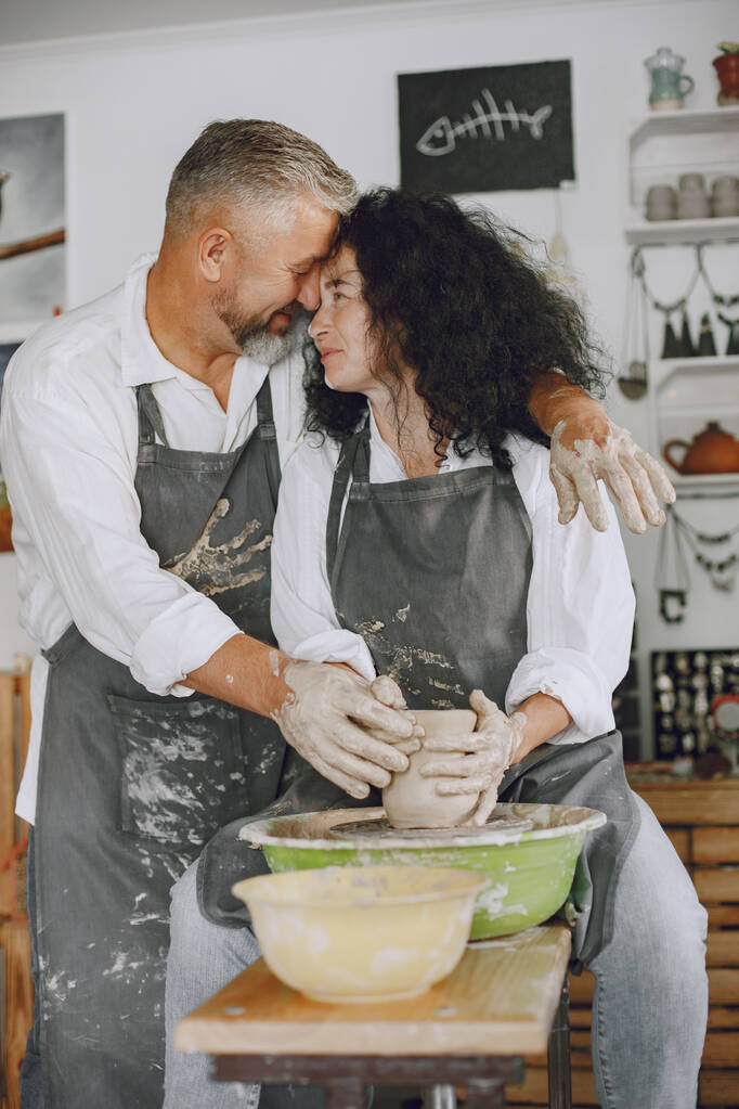 Trabalho criativo mútuo. Adulto casal elegante em roupas casuais e aventais. Pessoas criando uma tigela em uma roda de cerâmica em um estúdio de argila. - Foto, Imagem