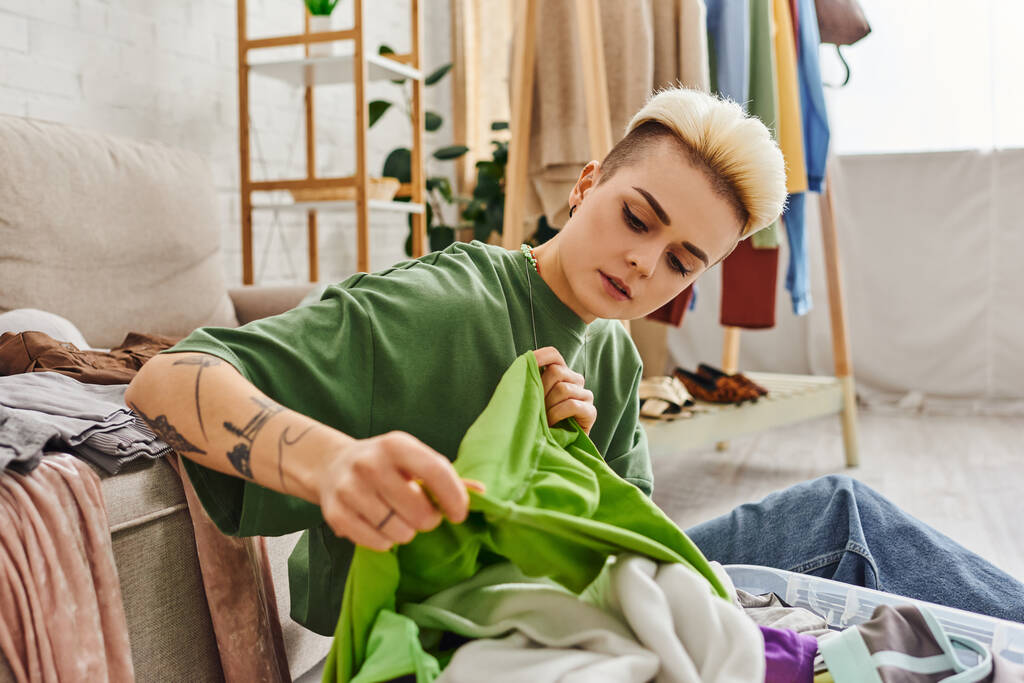 молодая, стильная и татуированная женщина с зеленой одеждой в современной гостиной у себя дома, сортировка и разглаживание, устойчивая жизнь и внимательный концепция потребительства - Фото, изображение