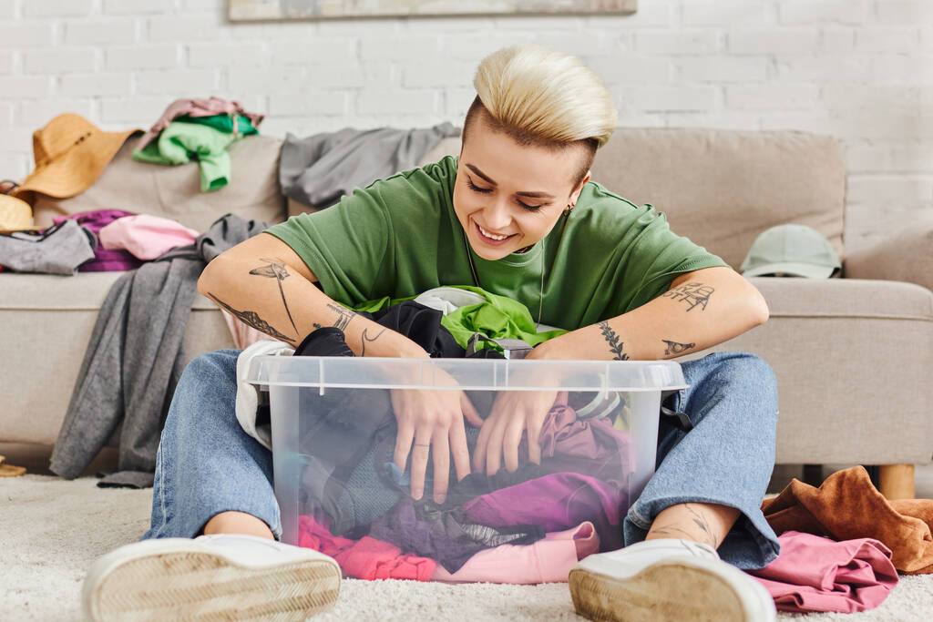 happy tattooed woman sitting on floor and sorting pre-loved items in plastic container near couch at home, trendy hairstyle, clothes decluttering, sustainable living and mindful consumerism concept - Photo, Image