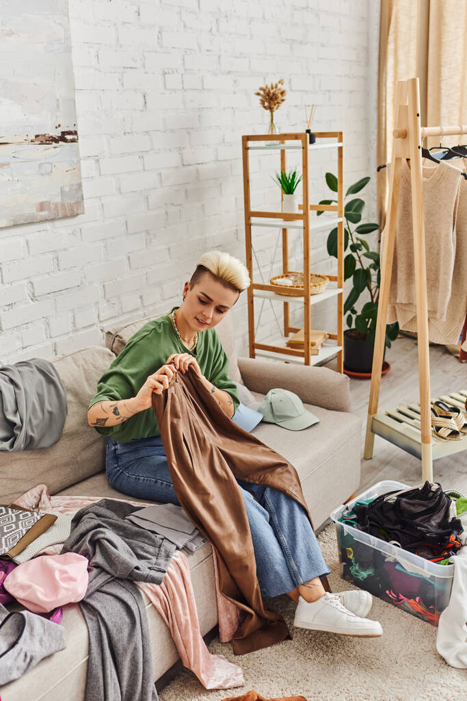 mladá tetovaná žena držící kožené kalhoty, zatímco sedí na gauči u oděvů, regálů a rostlin v moderním obývacím pokoji, odlučovací proces, udržitelný život a bdělý konzumní koncept - Fotografie, Obrázek