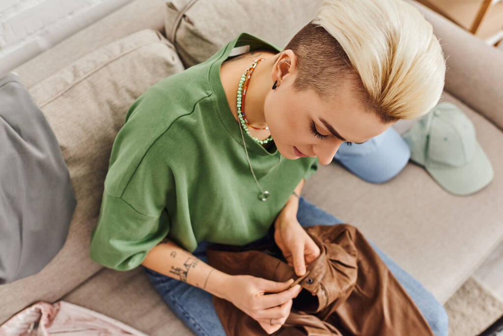 ylhäältä tyylikäs nainen trendikäs kampaus ja tatuointi istuu sohvalla lähellä vaatteita ja napittaa nahkahousut, decluttering prosessi, kestävä elävä ja tietoinen kulutuskäsite - Valokuva, kuva