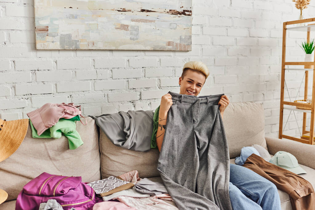 overjoyed tattooed woman holding grey pants and looking at camera near clothes on couch in living room, sorting wardrobe items, home decluttering, sustainable living and mindful consumerism concept - Photo, Image