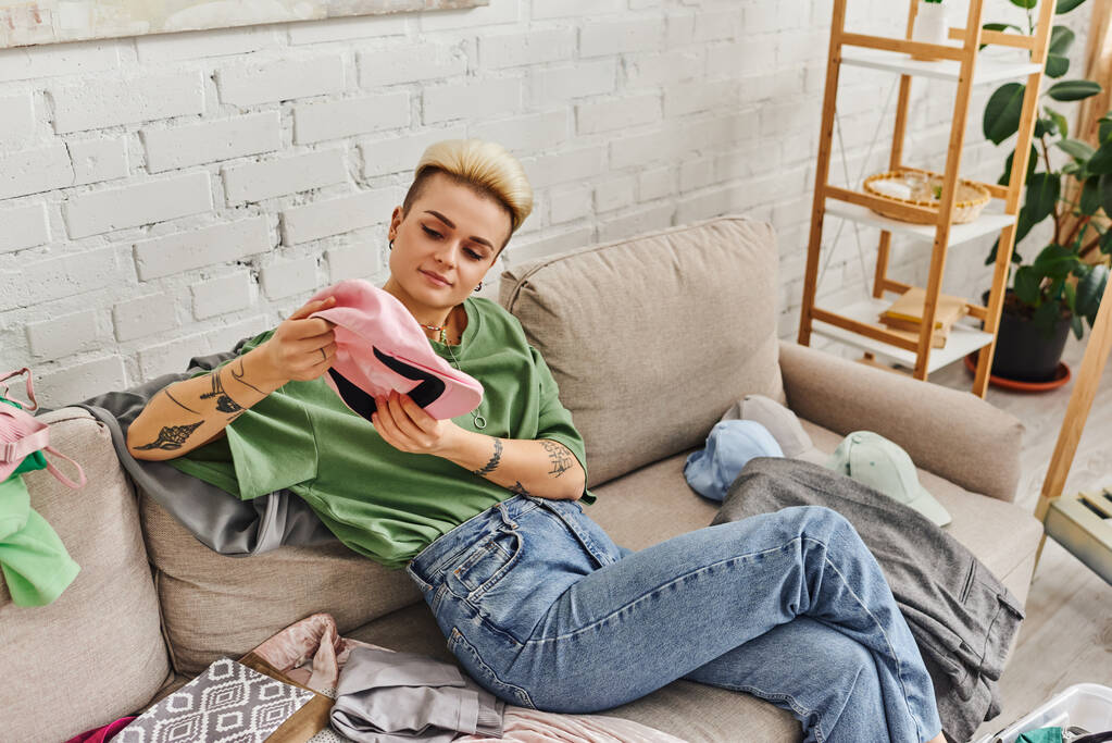 молодая женщина в повседневной одежде сидит на диване и держит шапку возле стойки и зеленое растение дома, модная прическа, татуировка, раздевание гардероба, устойчивая жизнь и внимательный концепция потребительского - Фото, изображение