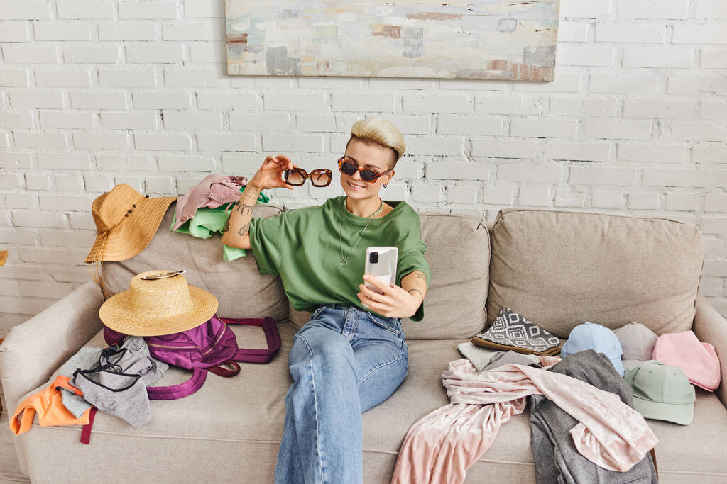pozytywna wytatuowana kobieta w luźnych ubraniach i stylowych okularach przeciwsłonecznych robiąca selfie na kanapie w pobliżu elementów garderoby do wymiany online, zrównoważonego życia i świadomego konsumpcjonizmu - Zdjęcie, obraz