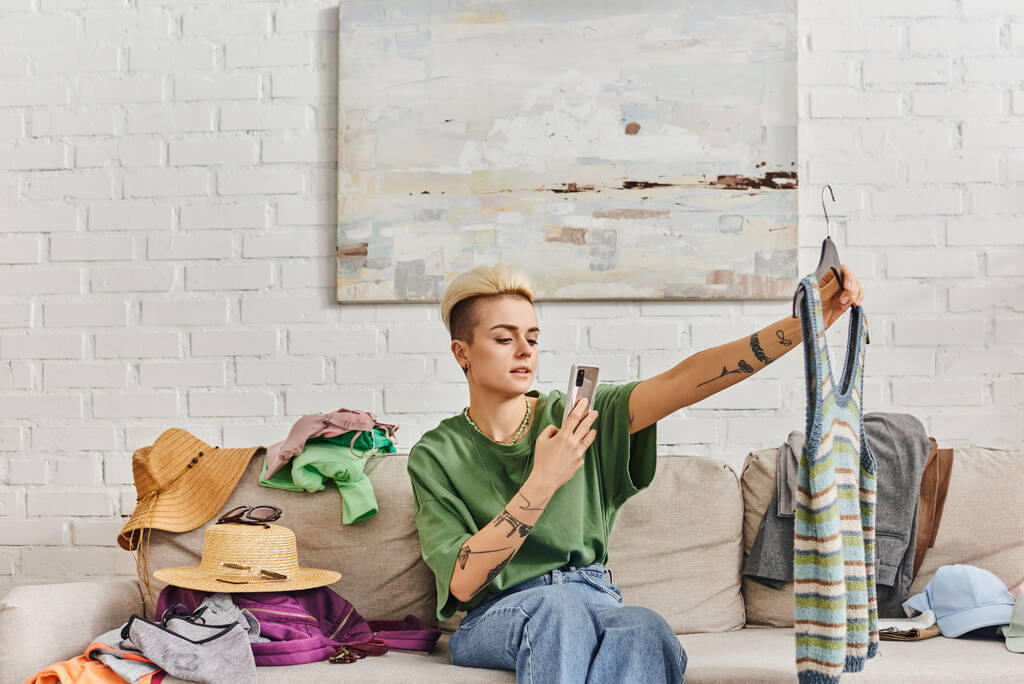 Tätowierte Frau mit Smartphone fotografiert gestrickte Tanktop auf Couch in der Nähe von Strohhüten und Kleidung, Online-Austausch, Kreislaufwirtschaft, nachhaltiges Leben und achtsames Konsumkonzept - Foto, Bild