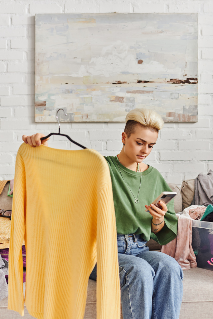 młoda, wytatuowana kobieta trzymająca żółty sweter i patrząca na telefon komórkowy siedząc na kanapie w pobliżu ubrań, sortując ubrania, wymieniając się online, zrównoważonym życiem i świadomym konsumpcjonizmem - Zdjęcie, obraz