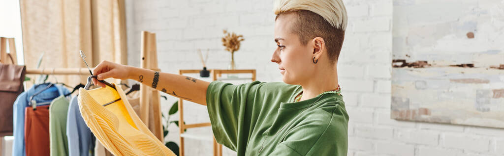 clothing sorting, side view of young tattooed woman with trendy hairstyle standing with yellow jumper near rack with garments on hangers, sustainable living and mindful consumerism concept, banner - Photo, Image