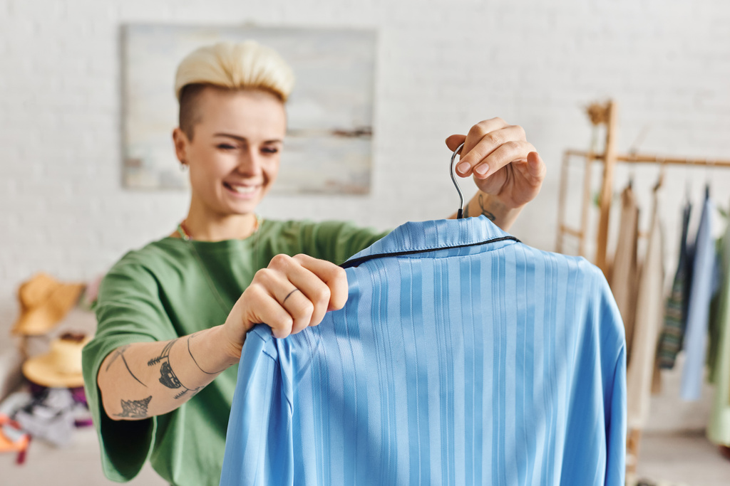 spaarzame kleding, kleding sorteren, tevreden getatoeëerde vrouw met hanger met blauwe pyjama in de woonkamer thuis op wazige achtergrond, duurzaam leven en bewust consumentisme concept - Foto, afbeelding