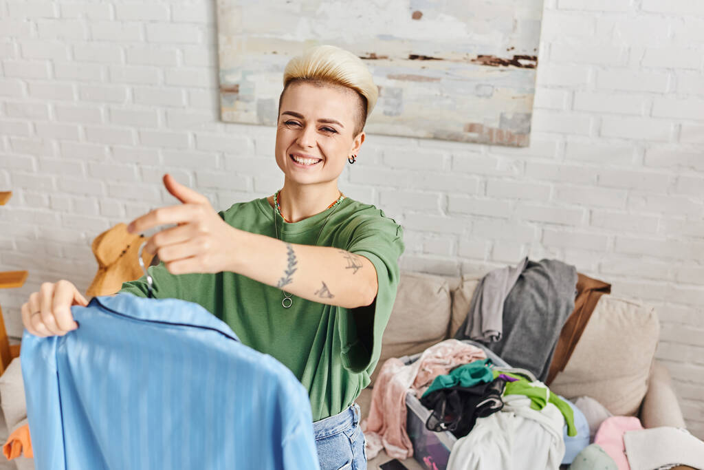 sorting clothing at home, young and joyful woman with tattoo and trendy hairstyle standing with blue pajamas near rack with wardrobe items at home, sustainable living and mindful consumerism concept - Photo, Image