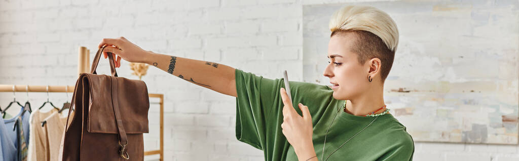 donna tatuata con acconciatura alla moda e smartphone che scatta foto di borsa in pelle vicino al rack con abiti stile casual, swap online, concetto di vita sostenibile e consumismo consapevole, banner - Foto, immagini
