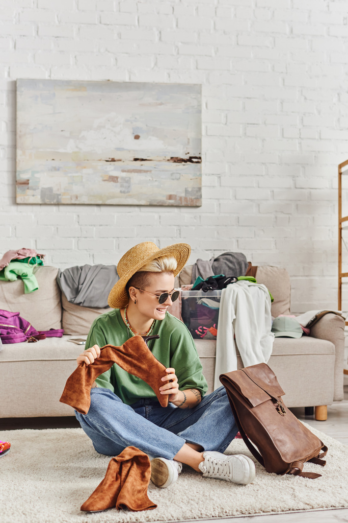 mujer sonriente y de moda en sombrero de paja y gafas de sol sentado en el suelo con bolsa de cuero y botas de gamuza cerca de sofá en la sala de estar, moda sostenible y concepto de consumismo consciente - Foto, imagen