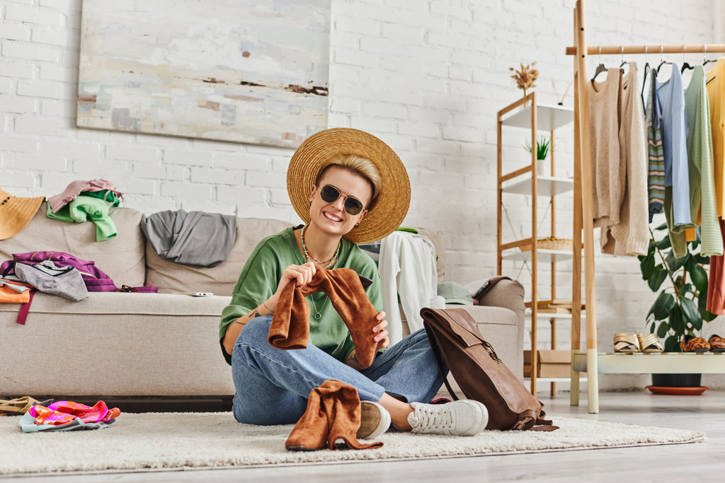 сортировка одежды, татуированная женщина в соломенной шляпе и солнцезащитных очках, смотрящая на камеру рядом замшевые сапоги и кожаную сумку на полу дома, устойчивая мода и внимательный концепт потребительства - Фото, изображение