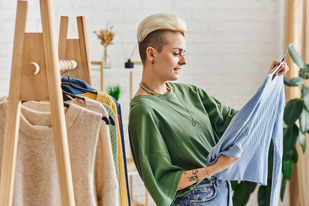 kleding sorteren, zijaanzicht van trendy en getatoeëerde vrouw kijken naar blauw vest in de buurt rek met garderobe items op hangers, duurzame mode en bewust consumentisme concept - Foto, afbeelding