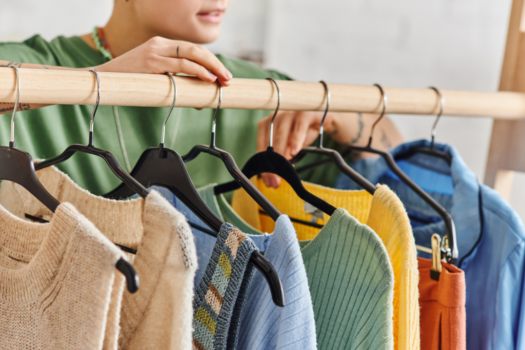 μερική άποψη της νεαρής γυναίκας στέκεται κοντά rack με μοντέρνα και πολύχρωμα casual ρούχα σε κρεμάστρες στο σαλόνι στο σπίτι, thrift κατάστημα βρίσκει, βιώσιμη μόδα και προσεκτική έννοια καταναλωτισμού - Φωτογραφία, εικόνα