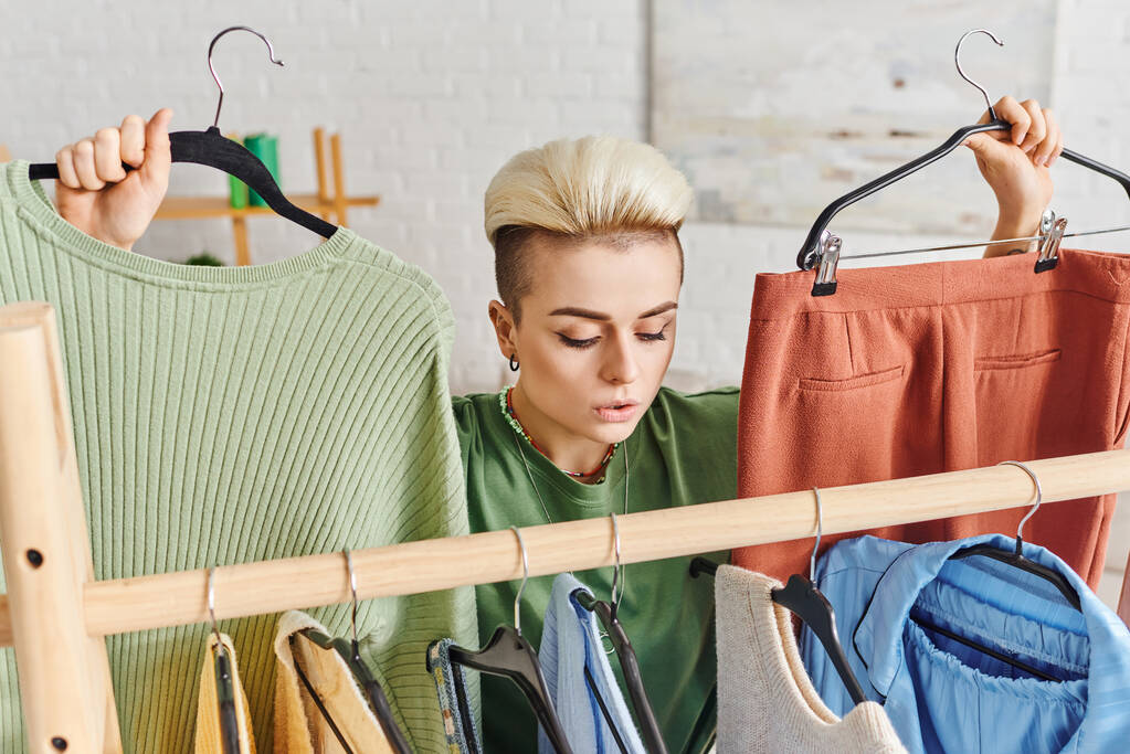 вдумчивая женщина с модной прической, держащая вешалки с брюками и свитер возле стойки с повседневной одеждой, домашнее раздевание, устойчивая мода и внимательный концепция потребительского - Фото, изображение