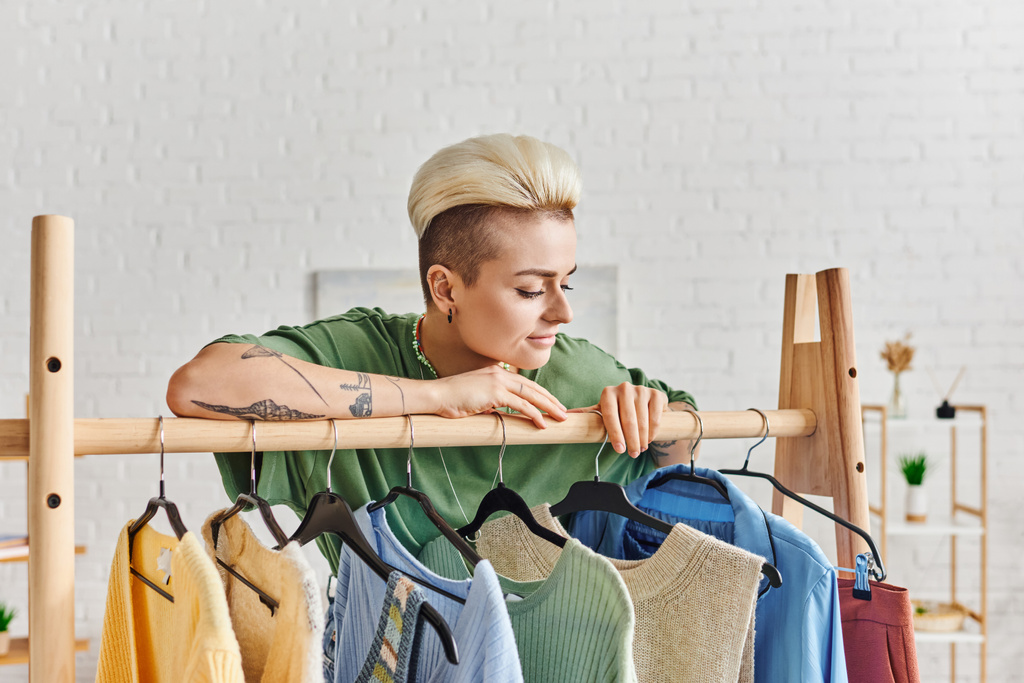 pozytywna i wytatuowana kobieta z modną fryzurą opierająca się na stojaku i patrząca na swobodną odzież na wieszakach w domu, przedkochane przedmioty, zrównoważoną modę i świadomą koncepcję konsumpcjonizmu - Zdjęcie, obraz