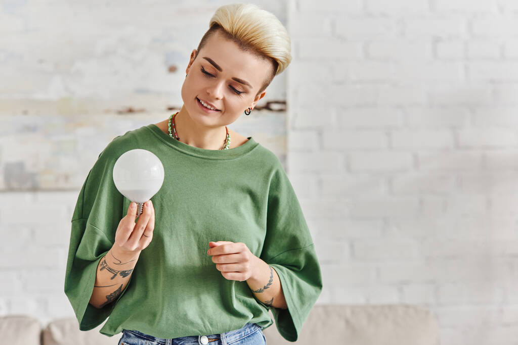 cheerful young woman in casual clothes looking at energy saving light bulb at home, trendy hairstyle, tattoo, positive emotion, sustainable lifestyle and environmentally conscious concept - Photo, Image