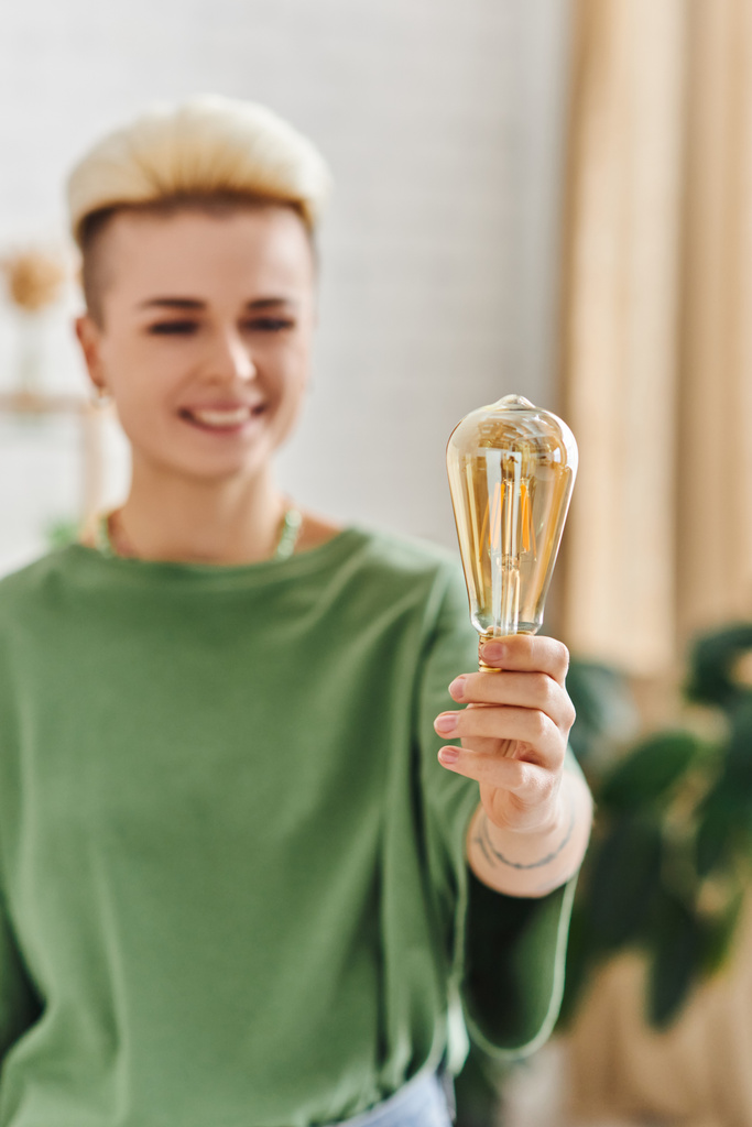 cheerful woman with trendy hairstyle, in casual clothes smiling and standing with energy saving light bulb at home on blurred background, sustainable lifestyle and environmentally conscious concept - Photo, Image