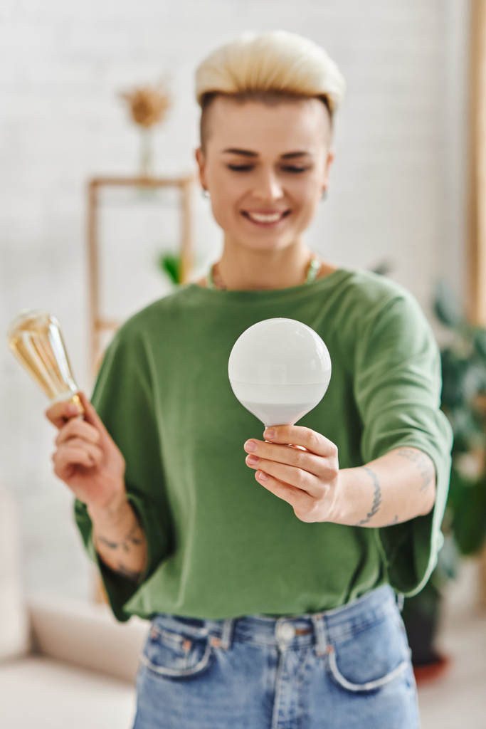 young and tattooed woman in casual clothes holding energy saving light bulbs and smiling in living room on blurred background,  sustainable lifestyle and environmentally conscious concept - Photo, Image