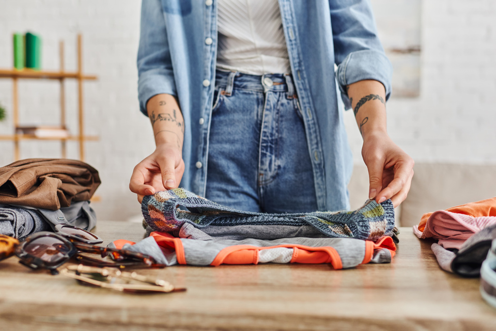 partial view of young and tattooed woman in casual clothes sorting wardrobe items on table in living room, exchange market, eco-friendly swaps, sustainable living and mindful consumerism concept - Photo, Image