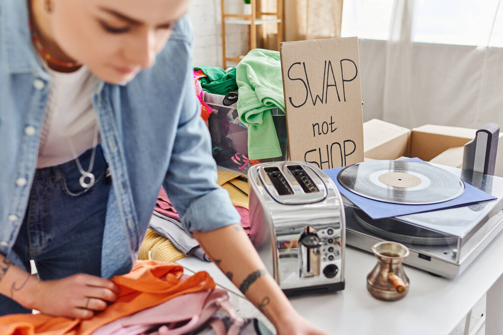 young tattooed woman sorting second-hand clothes near electric toaster, cezve, vinyl record player and swap not shop card, blurred foreground, sustainable living and circular economy concept - Photo, Image