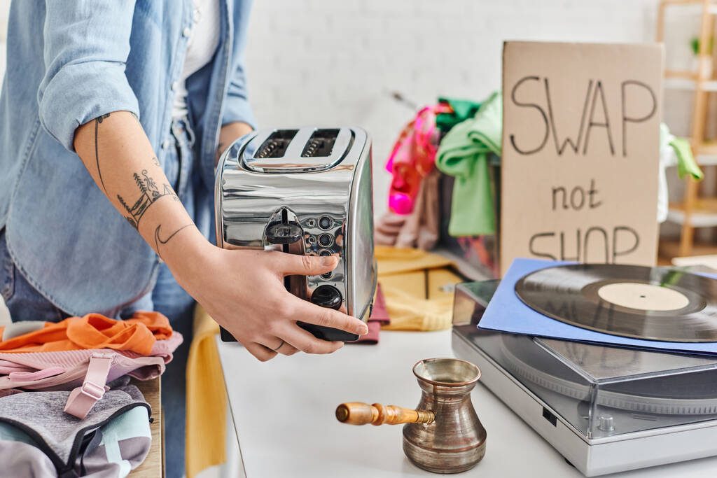 Teilbild einer jungen tätowierten Frau mit elektrischem Toaster in der Nähe von Plattenspieler, Cezve und Second-Hand-Klamotten während der Swap-Not-Shop-Veranstaltung, nachhaltigem Leben und Kreislaufwirtschaftskonzept - Foto, Bild