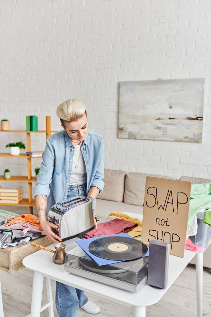 casually styled and tattooed woman holding electric toaster near vinyl record player, cezve, pre-loved items and swap not shop card in living room, sustainable living and circular economy concept - Photo, Image