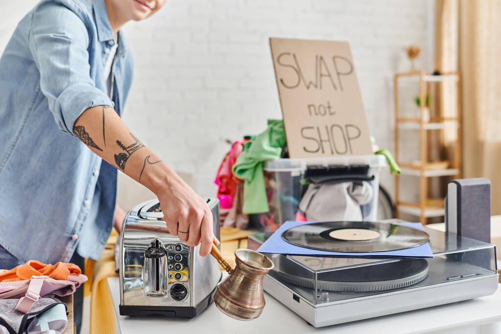 partial view of tattooed woman holding cezve near electric toaster, vinyl record player, clothes and card with swap not shop lettering at home, sustainable living and circular economy concept - Photo, Image